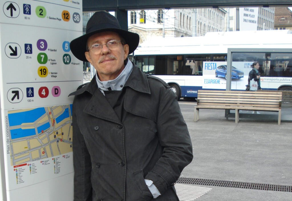 José Eduardo Heflinger Júnior encostado em uma placa do terminal de ônibus, na cidade de Genebra na Suíça, vestindo um sobretudo preto, chapéu preto e óculos de grau
